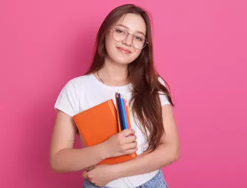 close-up-portrait-of-cute-young-woman-holding-textbook-and-colored-pencils-posing-in-studio-isolated-over-pink